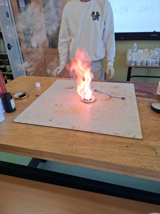 Osoba w laboratorium przeprowadza doświadczenie chemiczne z widocznym płomieniem na stole.