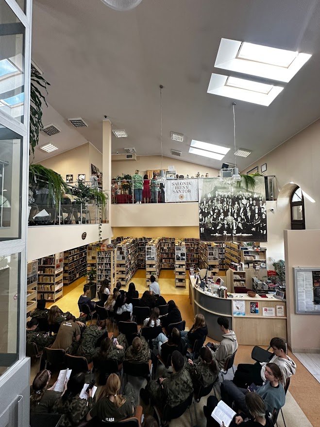Grupa młodzieży siedzi na krzesłach w bibliotece, trwa spotkanie lub prelekcja.
