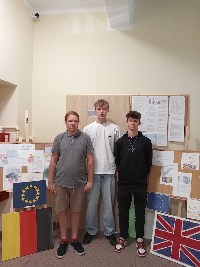Trzech chłopców stoi przed tablicami z pracami i flagami Unii Europejskiej, Niemiec i Wielkiej Brytanii.