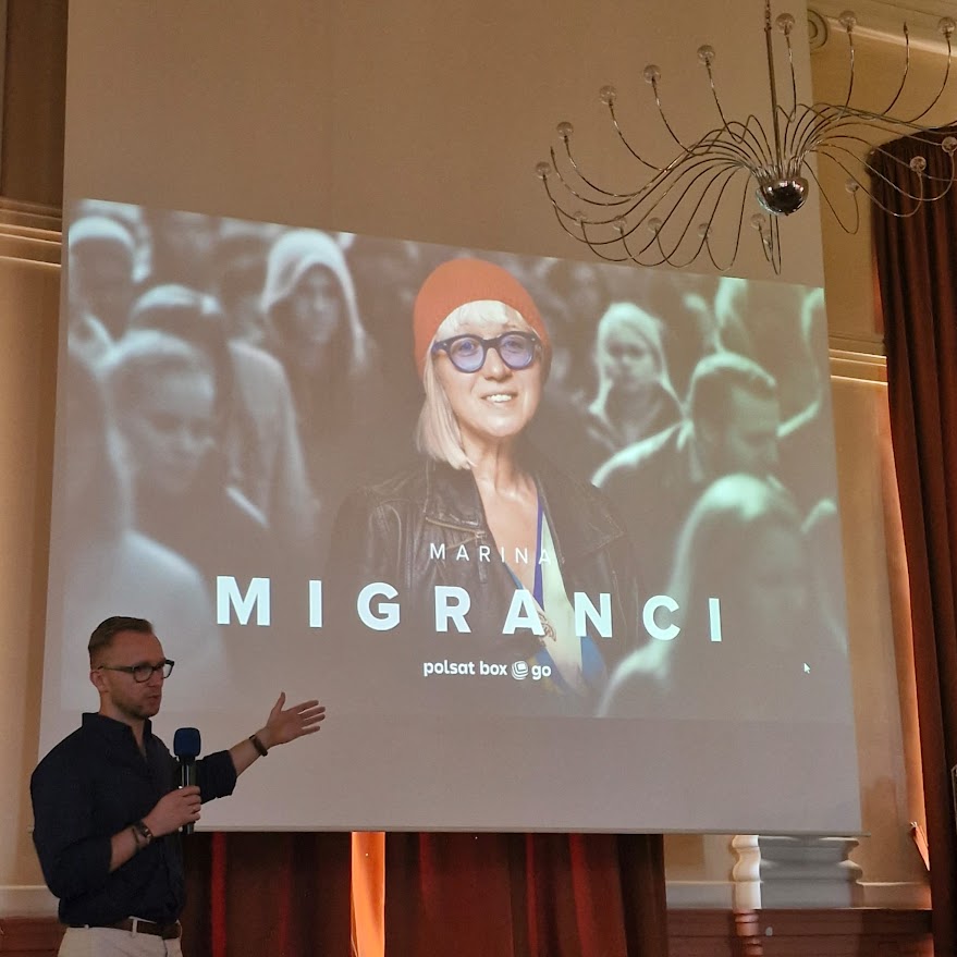Mężczyzna prezentuje slajd z plakatem filmu "Migranci" na dużym ekranie w sali.