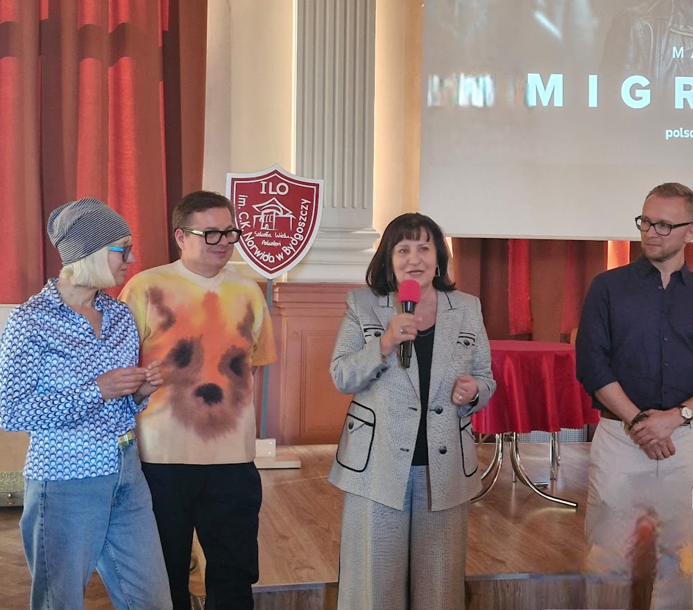 Cztery osoby stoją na scenie, kobieta w garniturze przemawia do mikrofonu przed publicznością.