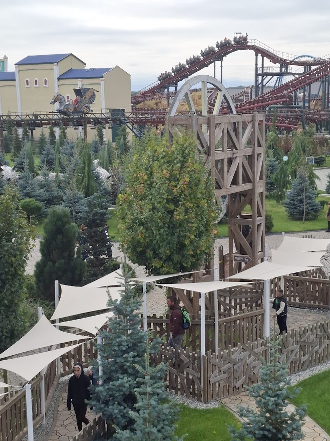 Ludzie spacerują w parku rozrywki, w tle widoczny rollercoaster i atrakcje tematyczne.
