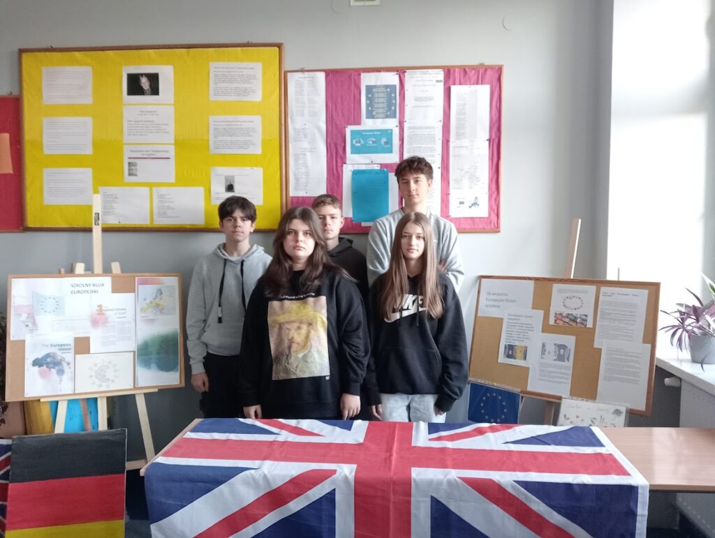 Pięcioro uczniów stoi przy stole z flagą Wielkiej Brytanii w sali szkolnej z planszami informacyjnymi.