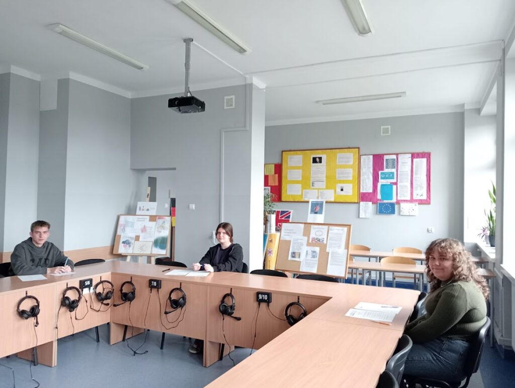 Trzech uczniów siedzi w sali językowej z słuchawkami na ławkach i materiałami edukacyjnymi.