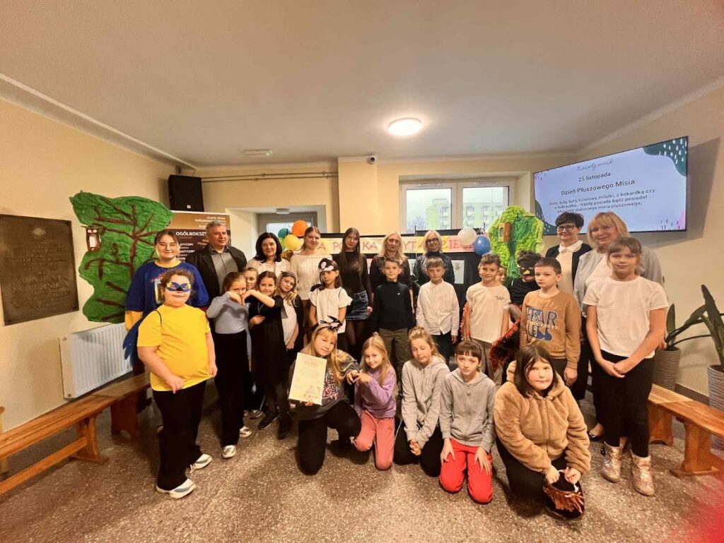 Grupa dzieci i dorosłych pozuje razem podczas Dnia Pluszowego Misia w szkole.