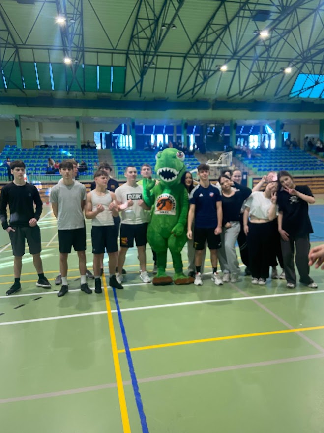 Grupa młodzieży pozuje do zdjęcia w hali sportowej z maskotką zielonego dinozaura.
