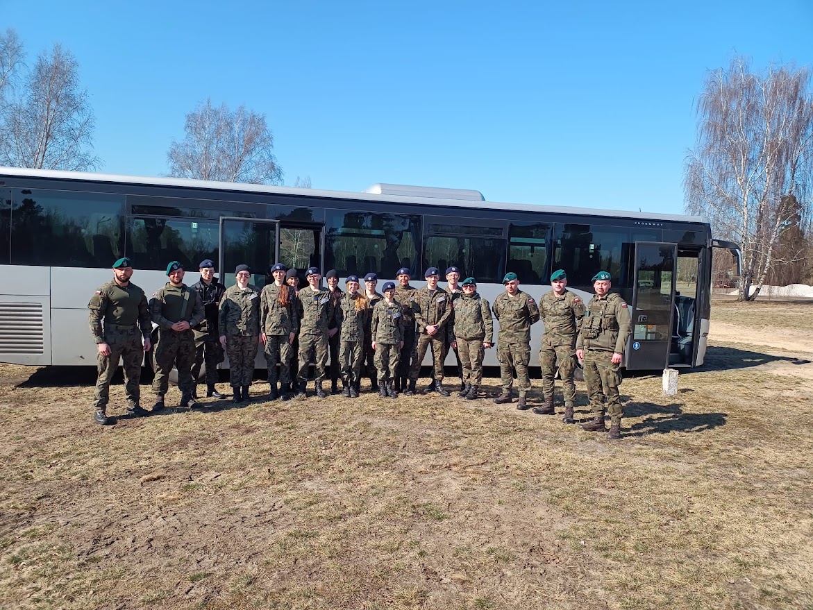 Grupa żołnierzy w mundurach stoi przed autobusem na otwartym terenie w słoneczny dzień.