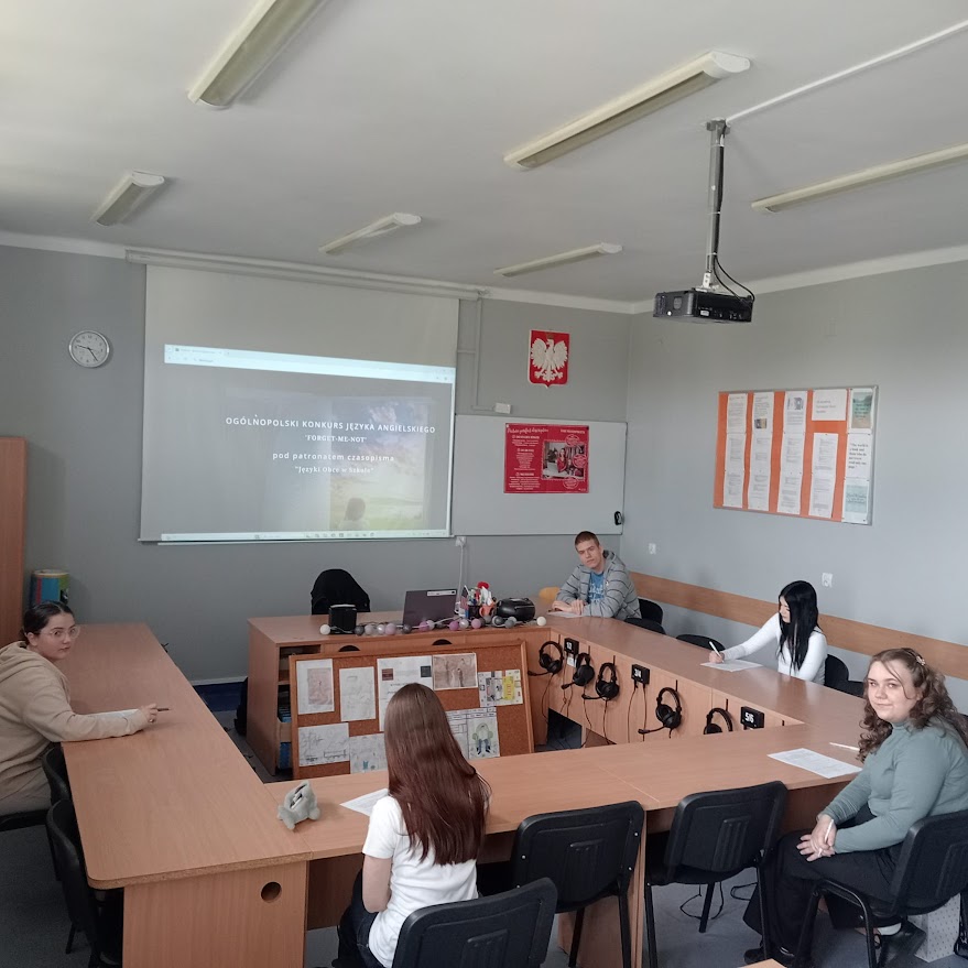 Kilka osób siedzi w sali lekcyjnej, na ekranie wyświetlony jest konkurs języka angielskiego.