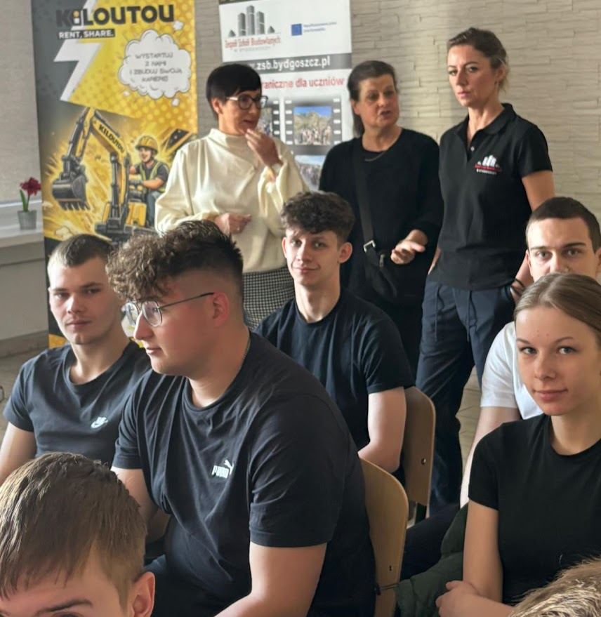 Grupa uczniów siedzi na krzesłach, za nimi stoją nauczycielki na tle banerów szkolnych.