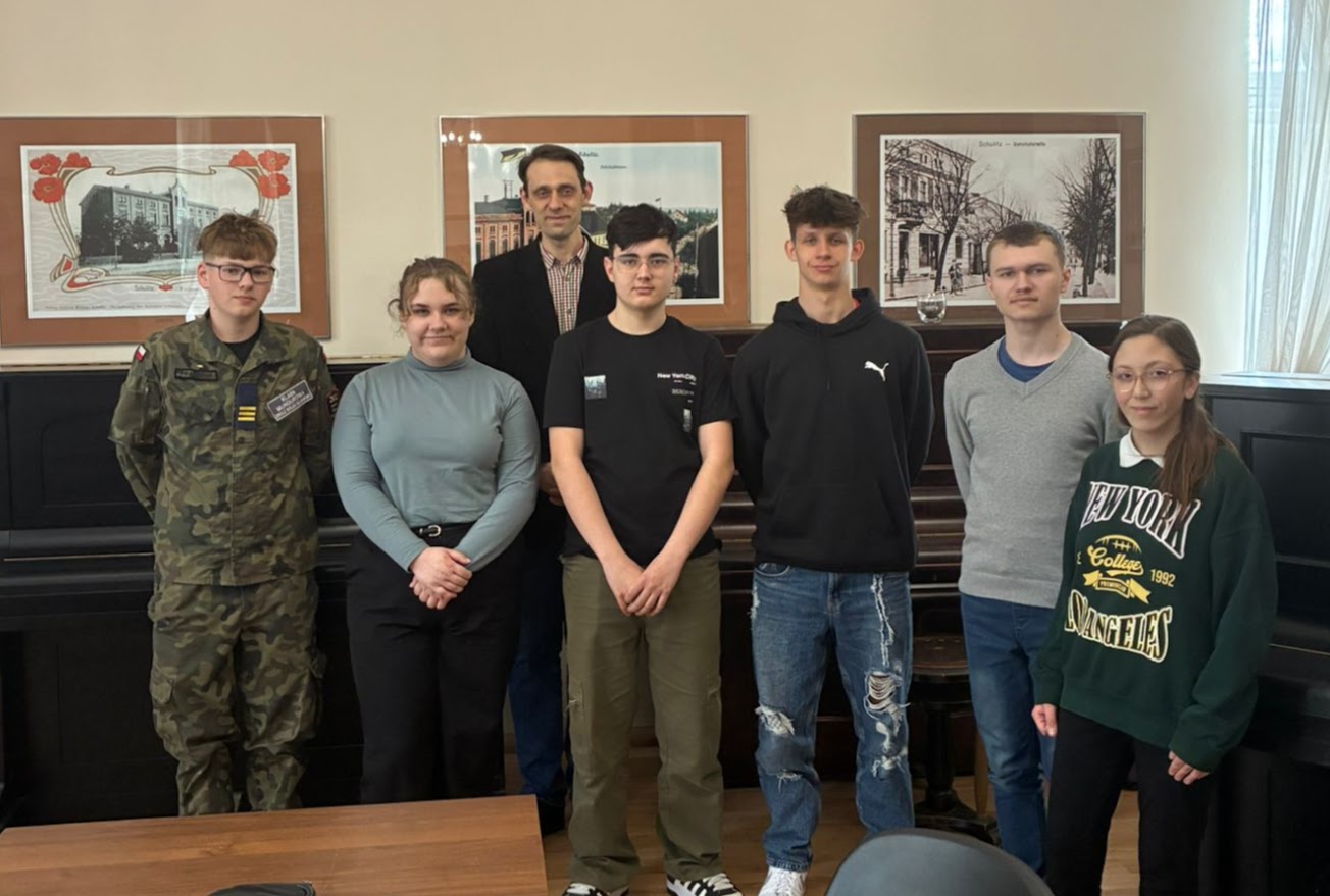 Grupa siedmiu osób pozuje do zdjęcia w sali z pianinem i obrazami na ścianach.