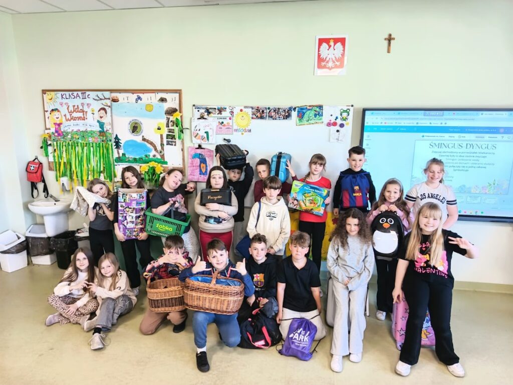 Grupa dzieci w klasie pozuje do zdjęcia, trzymając różne nietypowe torby zamiast plecaków.