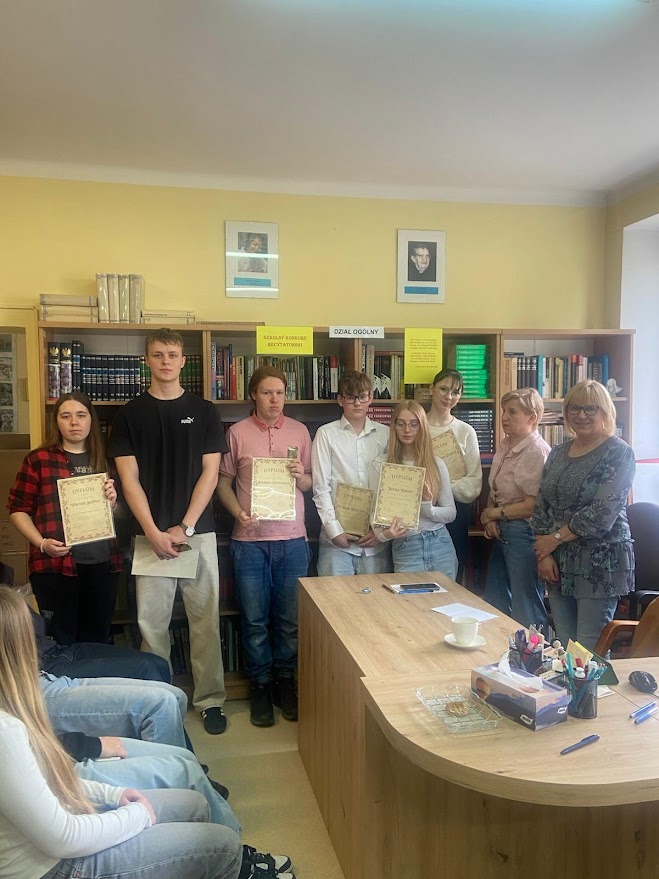 Grupa młodych osób i nauczycielek w bibliotece, kilka osób trzyma dyplomy za osiągnięcia.