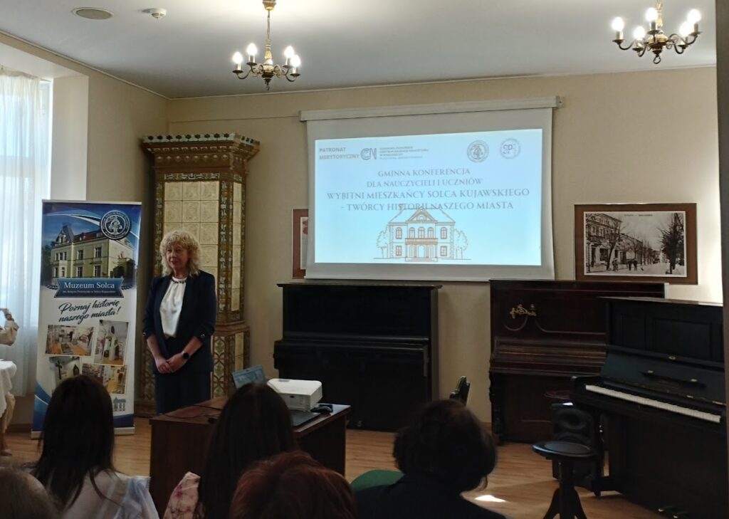 Kobieta przemawia podczas konferencji w muzeum, w tle ekran z prezentacją i pianino.