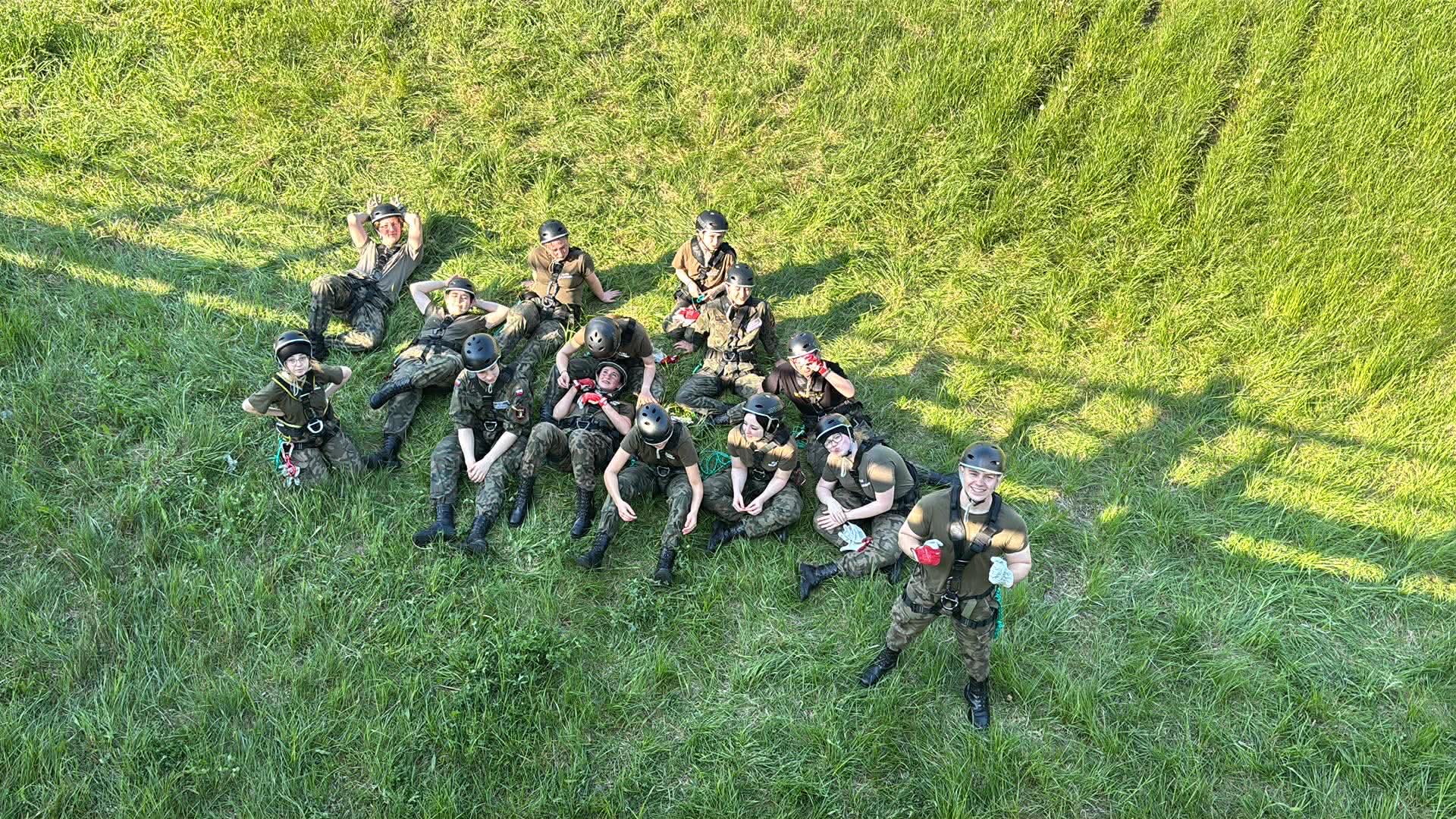 Grupa młodych osób w mundurach i kaskach siedzi i stoi na trawie, patrząc w górę.