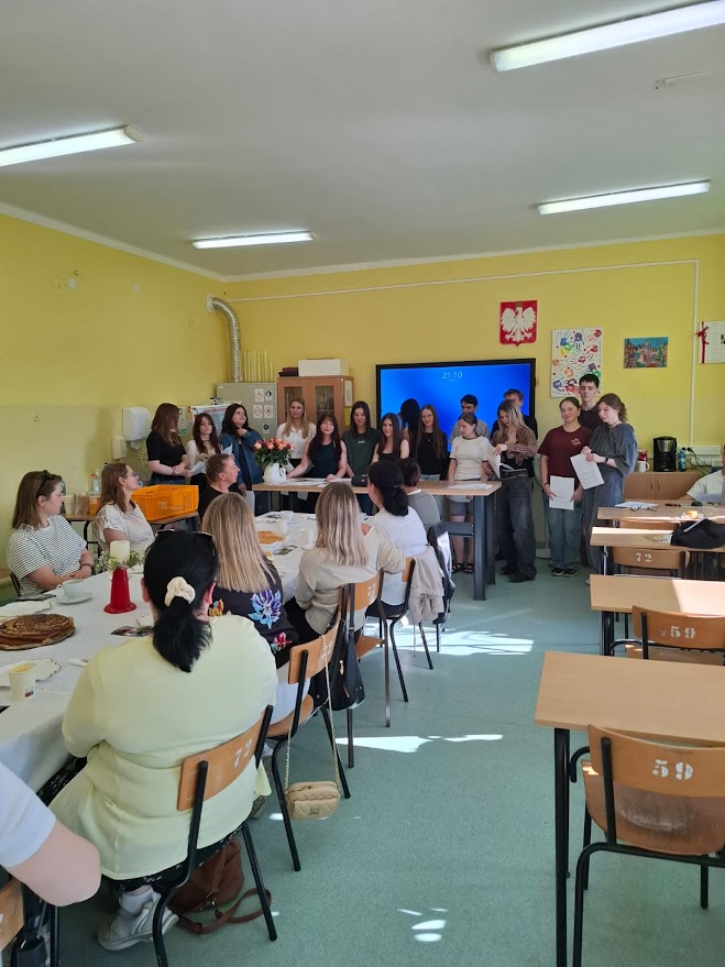 Grupa uczniów stoi przed klasą, pozostali siedzą przy stołach podczas szkolnej uroczystości.