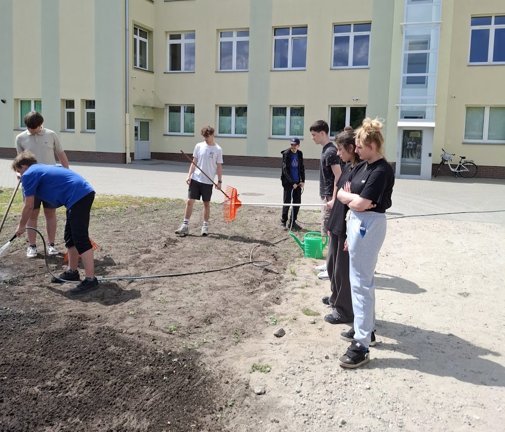 Grupa młodzieży pracuje w ogrodzie przed szkołą, używając grabi i konewki.
