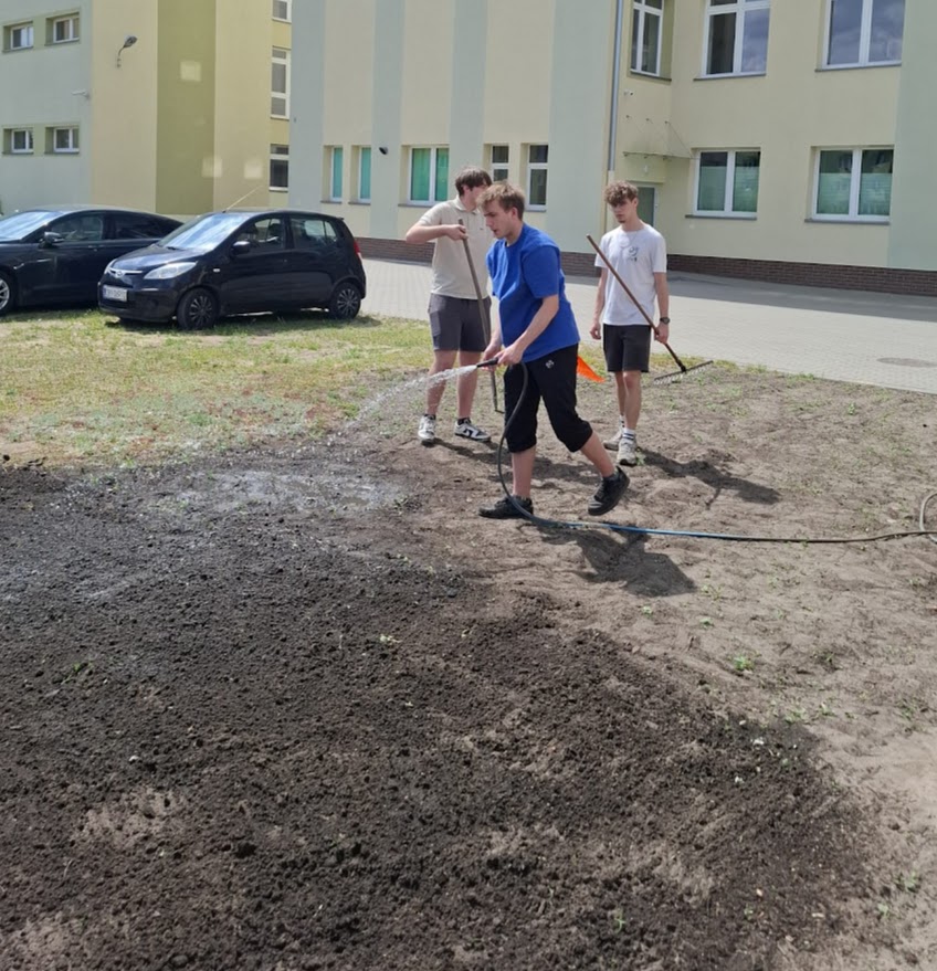 Trzech chłopców podlewa i grabi ziemię przed budynkiem szkolnym.