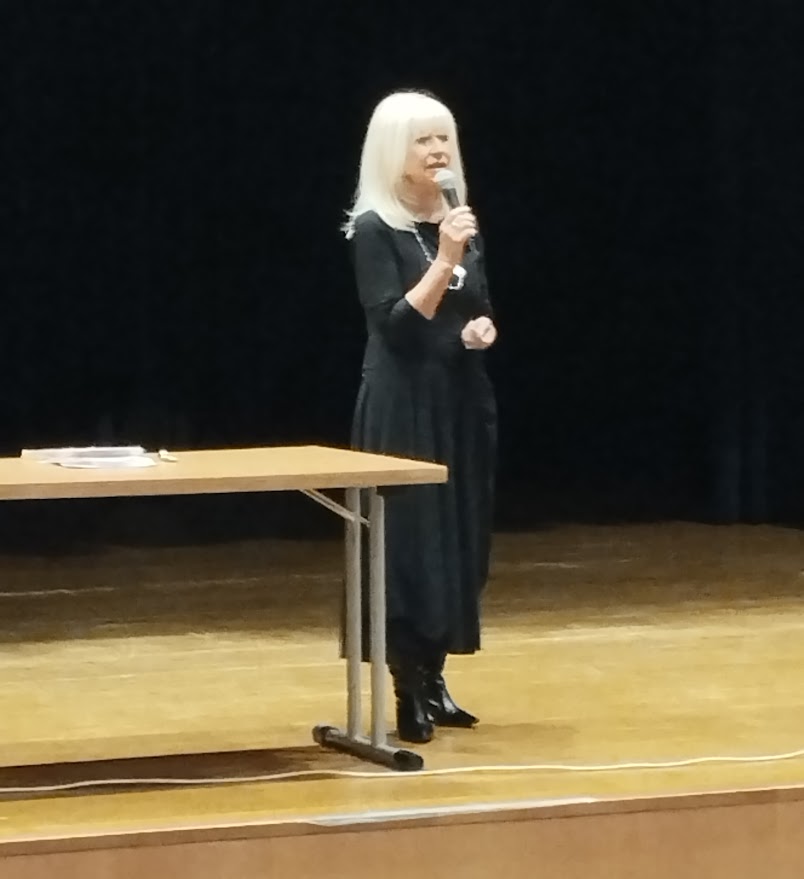 Kobieta w czarnej sukience stoi na scenie i przemawia do mikrofonu obok stołu.